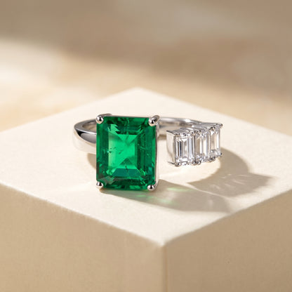 Open ring with a Colombian green emerald and three diamond side stones on a beige background; all gemstones are emerald cut