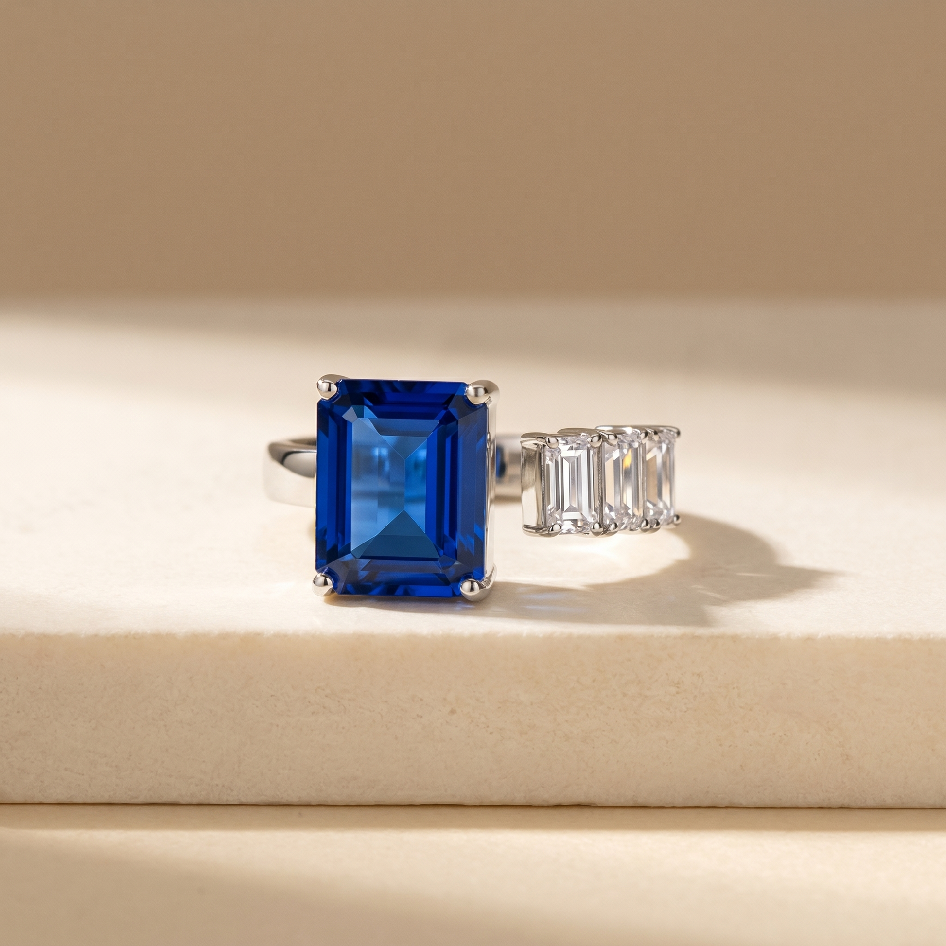 Open ring with a blue sapphire and three diamond side stones on a beige background; all gemstones are emerald cut