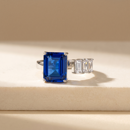 Open ring with a blue sapphire and three diamond side stones on a beige background; all gemstones are emerald cut