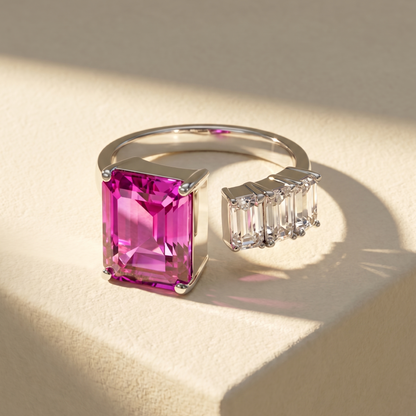Open ring with a vivid pink sapphire and three diamond side stones on a beige background; all gemstones are emerald cut