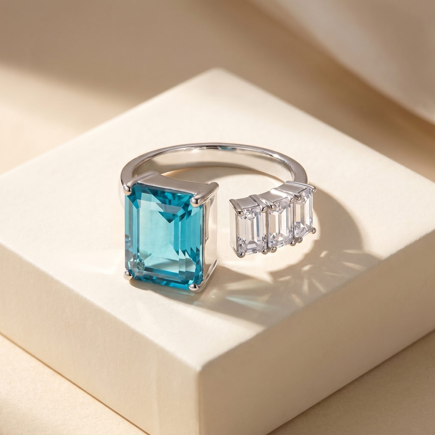 Open ring with a teal blue spinel and three diamond side stones on a beige background; all gemstones are emerald cut