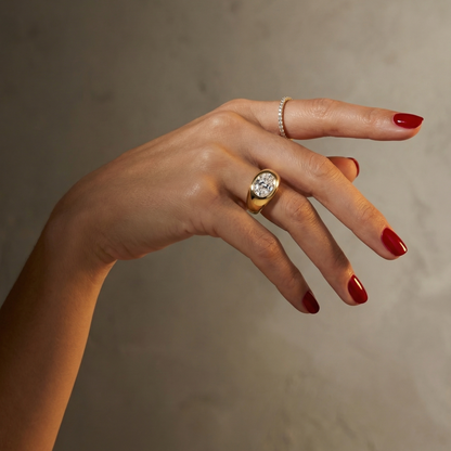 Hand wearing a statement, gold signet ring with a large oval diamond laterally set; hand shown on a plain background