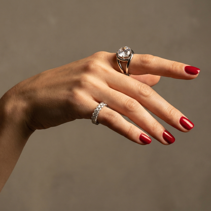 Hand with red nail polish wearing two diamond rings - one cocktail, statement, large oval diamond ring and one oval eternity ring worn as a pinky ring; hand is shown on a neutral background