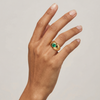 Hand wearing a gold signet style ring with a green Colombian Emerald round gemstone on a neutral background