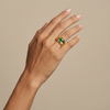 Hand wearing a gold signet style ring with a green Colombian Emerald round gemstone on a beige background