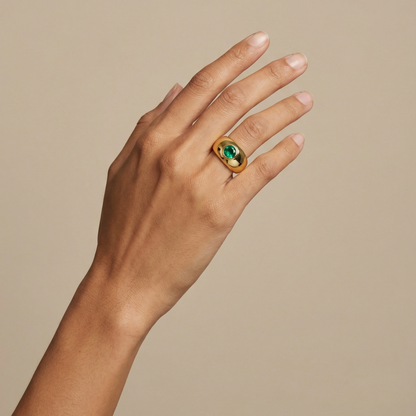 Hand wearing a gold signet style ring with a green Colombian Emerald round gemstone on a beige background