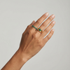 Hand wearing a gold signet style ring with a green Colombian Emerald round gemstone and an eternity ring in emerald on a neutral background
