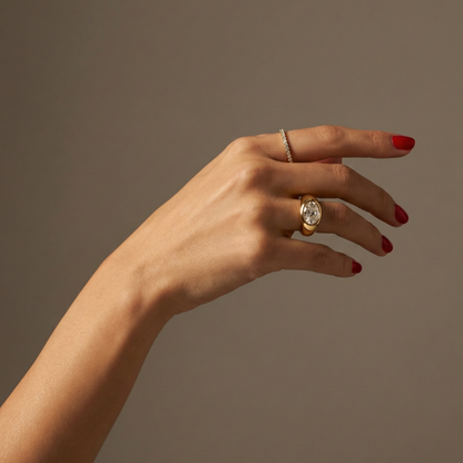 Hand wearing a statement, gold signet ring with a large oval diamond laterally set; hand shown on a plain background