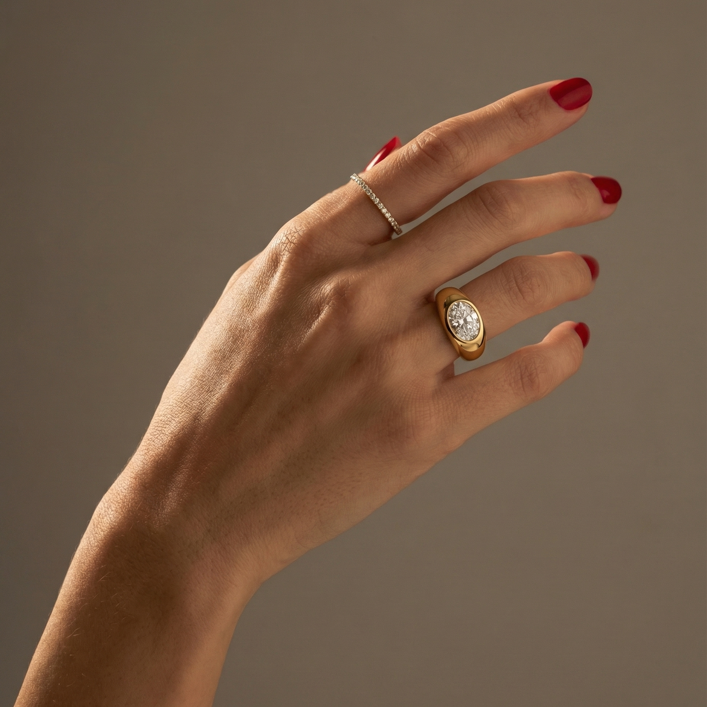 Hand wearing a statement, gold signet ring with a large oval diamond laterally set; hand shown on a plain background